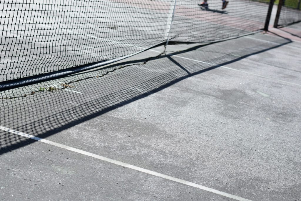 Outdoor tennis court viewed across a black net in bright sunlight