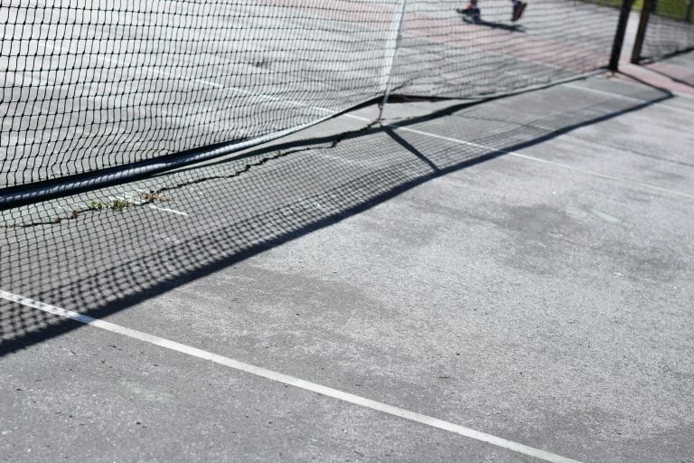 Outdoor tennis court viewed across a black net in bright sunlight