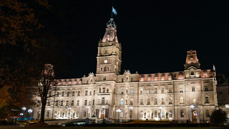 The illuminated Québec Parliament Building at night with a Québec flag flying above the central tower.