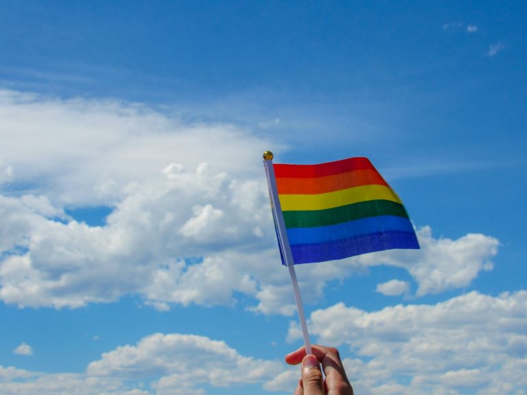 A small rainbow Pride flag held against a blue sky.