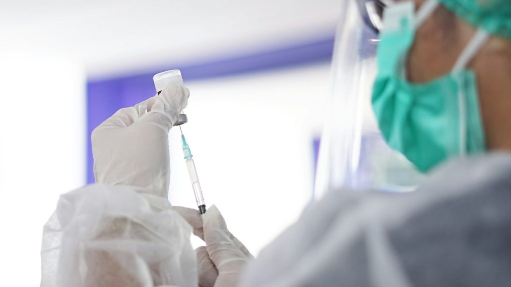 A health worker wearing protective equipment prepares a syringe from an unlabelled vial.